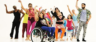 In this photo, over ten people dressed in yellow, red, blue, and other colors stand on a stage with a white dance floor. They are of different gender, age, and body shape, but all are doing a similar dance posture, standing on the right leg with the left leg pointing to the floor, stretching their left hand forward, the right hand up, and tilting the head slightly to the left.