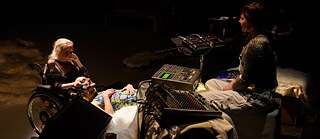 This picture shows a relaxed moment in the performance when sound artist Kim sits on the right side behind her mixing consoles, and audio equipment, with closed eyes and her hands resting in her lap. On the left side sits Lucy in a wheelchair, and Pawel is resting with his head on Lucy’s lap and knees, while his torso and legs are lying on a white flokati rug on a platform.