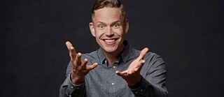 This picture is a portrait of the deaf Finnish rapper Signmark who is smiling brightly at the camera with his hands wide open in the front. He is standing in a studio with a dark background, wearing a gray button up shirt and a pair of black jeans with a brown belt. 