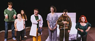  Seven people are on a theater stage. Behind them, there is a big bed. On the left is a young man in jeans and a sports jacket, followed by a girl with long hair and glasses, and a man with a white beard. On his right side are two women, one in a doctor’s jacket and the other in a worker uniform. Next to them are a man with sunglasses holding a guide stick, and a woman with red hair.