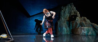  In this staged photo, Julia Häusermann is playing the role of the lead character, Frank. The woman has orange ponytail hair and is wearing a black dress with bright red trousers and white sneakers. The image is in dim blue and black, with the shadow of the heroine and a rockery in the background.