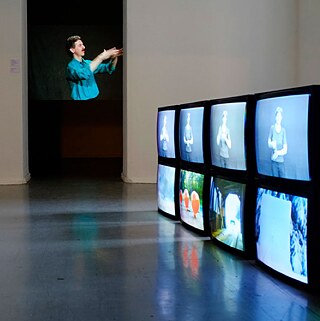 This photo shows an empty room in which eight CRT televisions are set up, showing different scenes. On the white wall directly opposite is a rectangular doorway. In the center of the doorway stands a man wearing a blue shirt and black trousers, with slightly white hair, holding his arms up to his eyes.