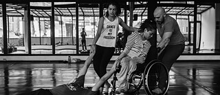 This black and white picture shows four people, two young women, and two men, in what looks like a dance studio. A young man is on the floor, and the two women, one in a wheelchair and one with no disabilities, are pulling him by his hands. The other man is pulling the women back. The T-shirt of the woman standing says: “Dance is my freedom”. 