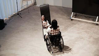 《生活多美好》现场演出图一：一位轮椅使用者女性演员正对着化妆镜中的自己，她身着菱格毛衣，黑色的中短发披在肩上。