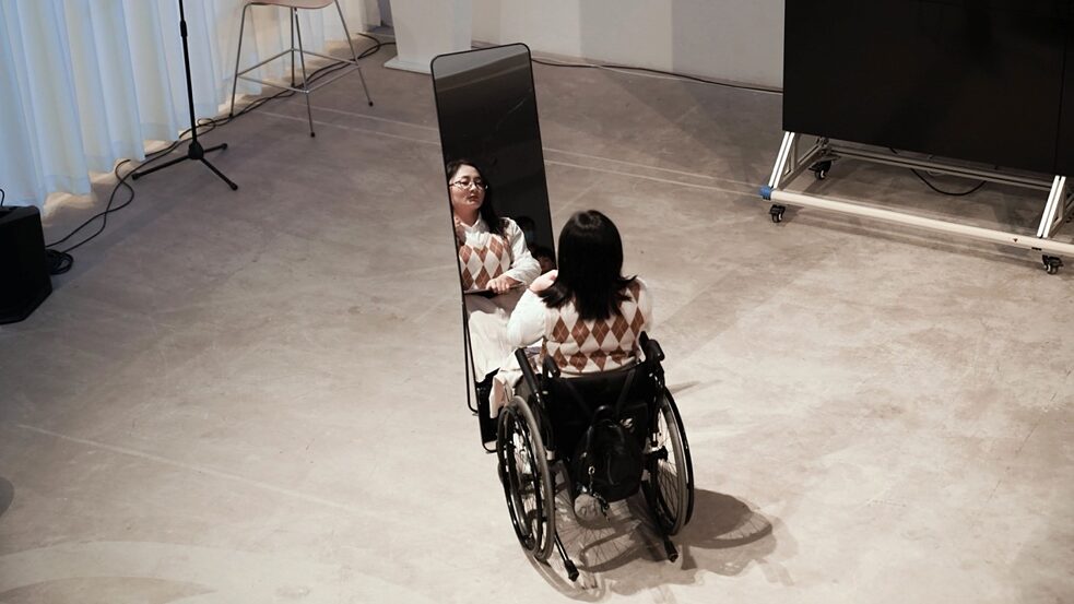 Figure no. 1 from the performance of What a Wonderful Life: A female wheelchair user looks at herself in the mirror. She is wearing a diamond-patterned sweater with black medium-length hair over her shoulders.