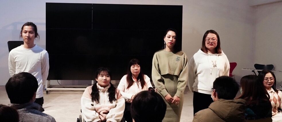 Figure no. 2 from the performance of What a Wonderful Life: Six actors stand in a row to make speeches. From left to right are an actor in a white T-shirt, two actresses in wheelchairs (the one on the left has two pigtails, wearing a thick pale pink coat, and the one on the right has long black hair, wearing a pale pink top), two actresses standing on the right (the one on the left has delicate makeup, wearing a camel-colored dress and the one on the right is wearing glasses and a white hoodie), and an actress in a wheelchair who also appears in Figure no. 1. This is the last scene where the actors are on stage together.