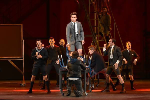 A photo of Spring Awakening, featuring a group of male actors dancing with their arms in the air and the male protagonist in the middle standing on a chair and facing forward